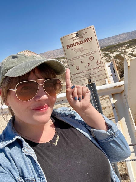 Photo of a woman with aviator sunglasses and a baseball cap pointing at a desert sign 