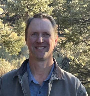 A man with brushed back brown hair wearing a blue shirt and a jacket